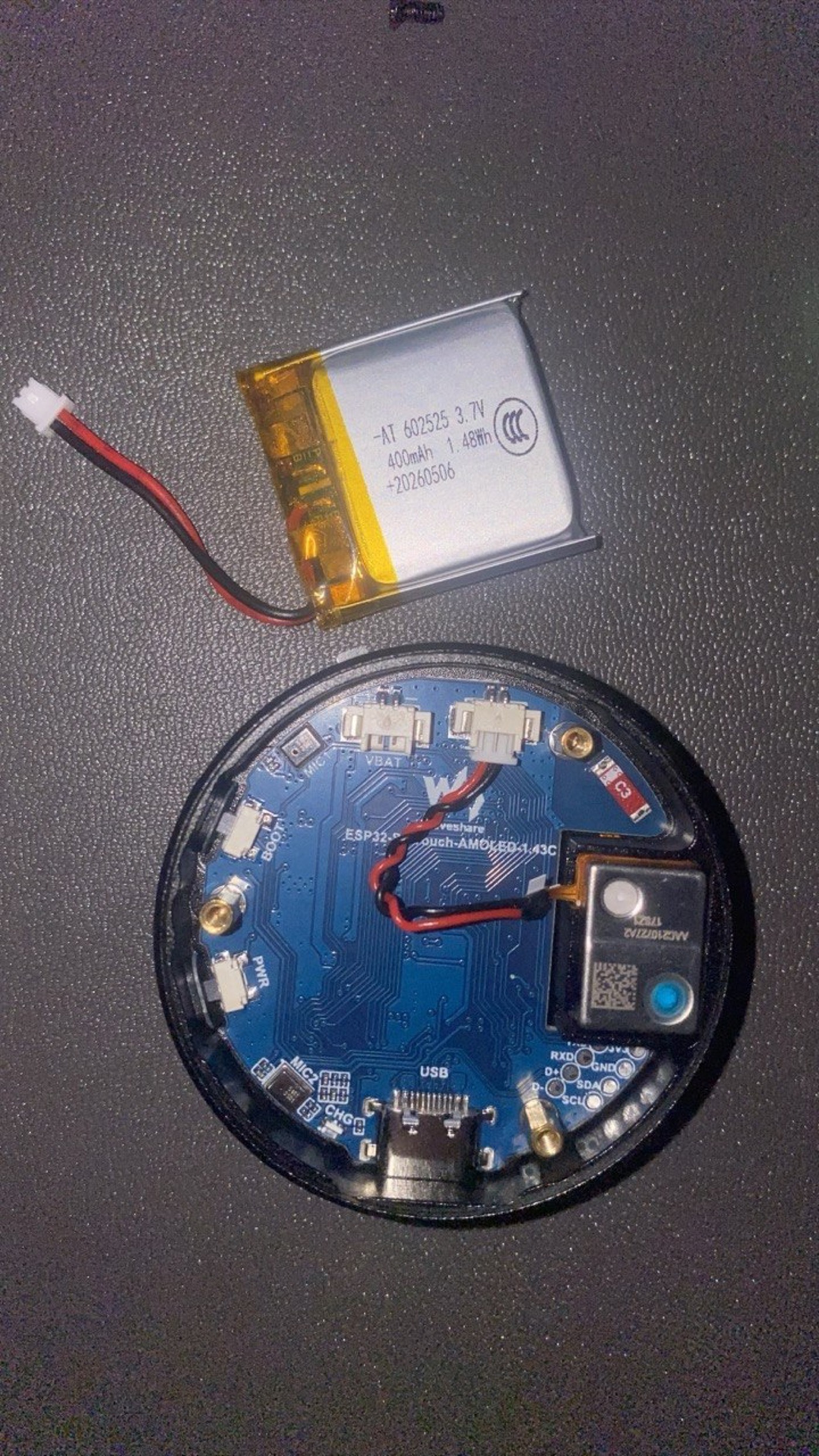 The board opened up — the round AMOLED, the LiPo cell, and the microphone that makes the voice side work